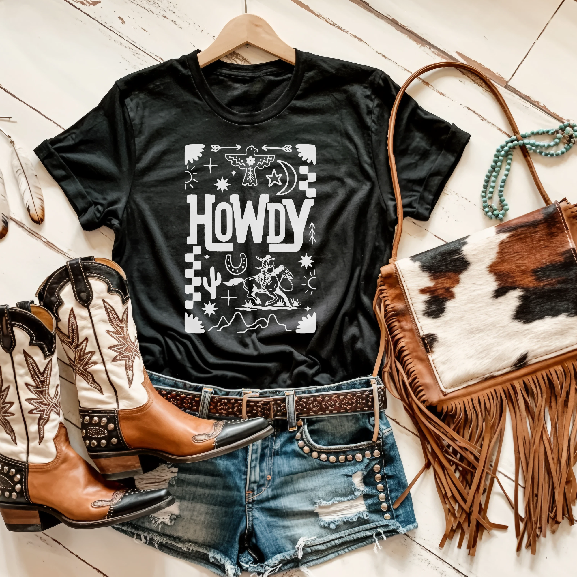 Black t-shirt with 'HOWDY' graphic, cowboy boots, denim shorts, and a fringe bag on a wooden floor.