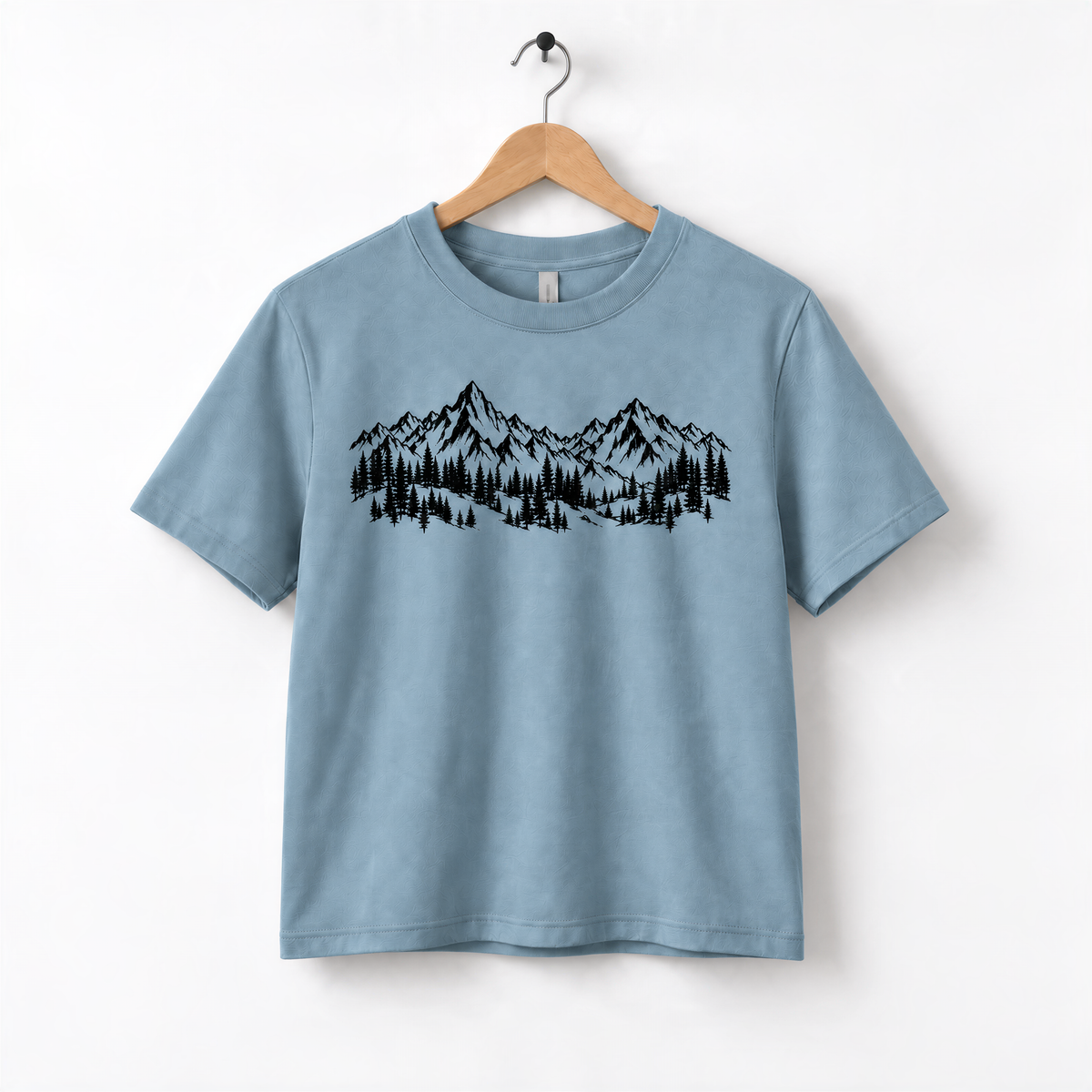 A light blue t-shirt with a graphic design of mountains and trees printed on it, hanging on a wooden hanger.