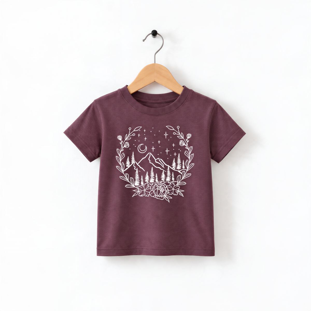 A burgundy-colored t-shirt with a white graphic design depicting a mountain landscape with trees, flowers, and stars.