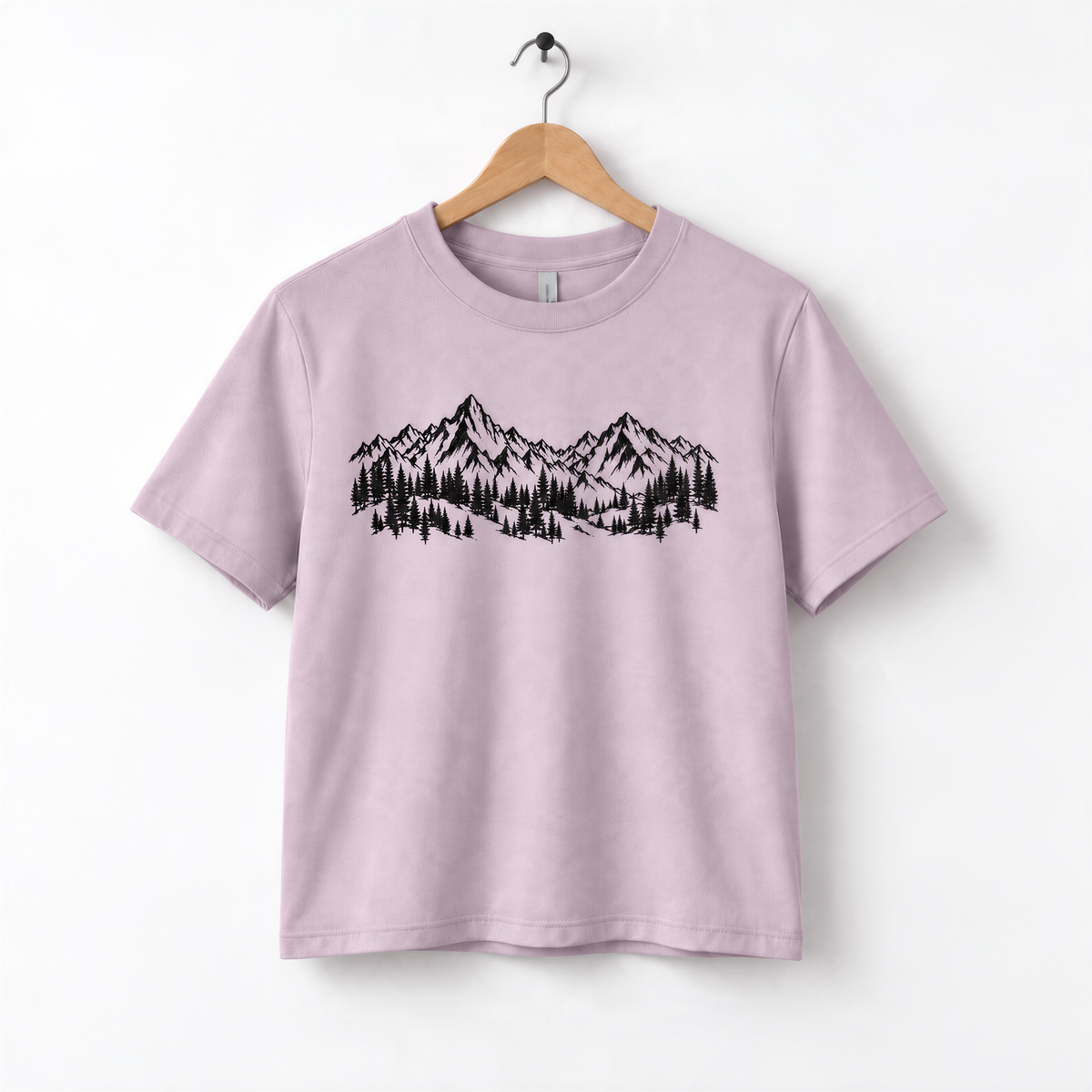 A light pink t-shirt with a graphic design of mountains and trees printed on it, hanging on a wooden hanger.