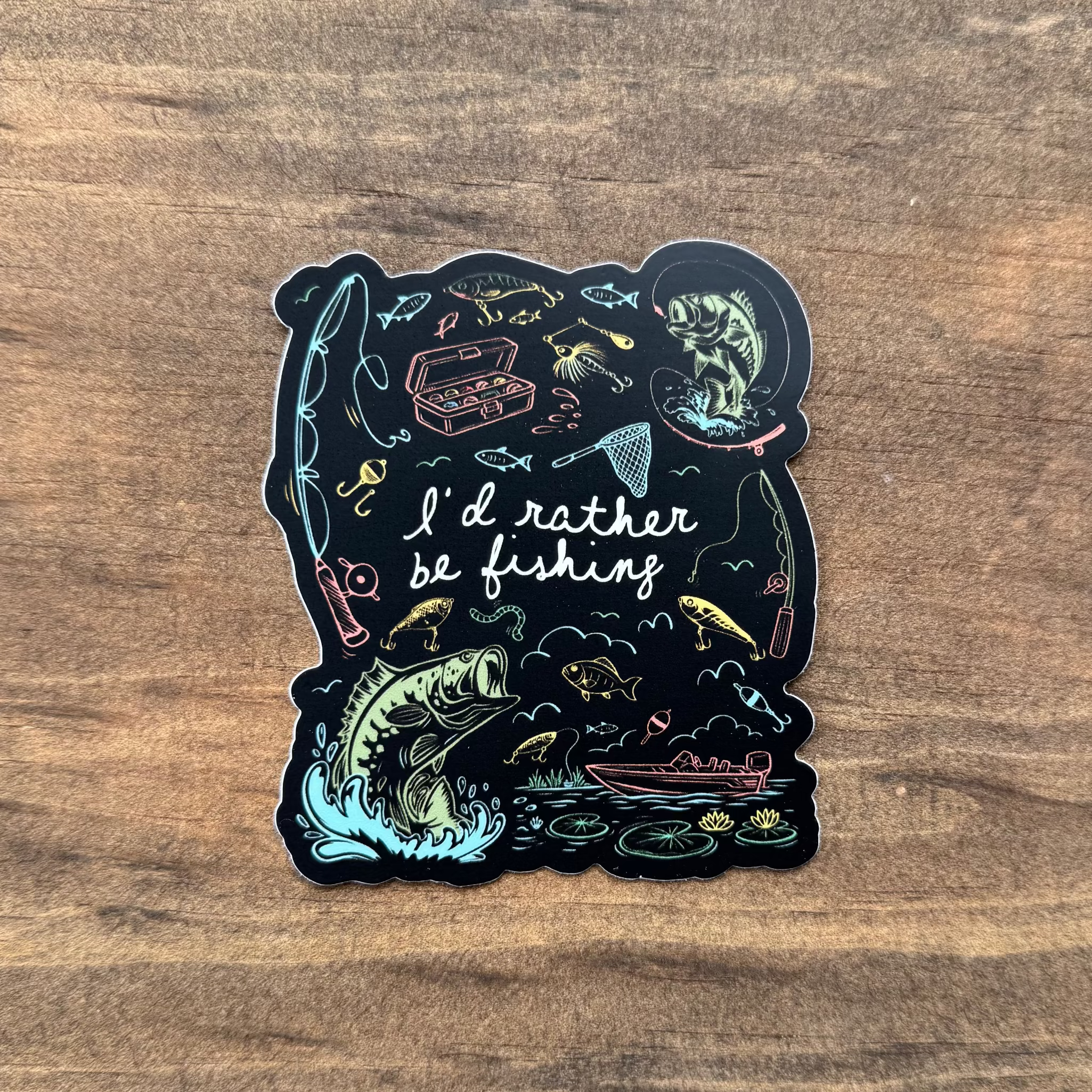 Sticker with fishing-themed design and 'I'd rather be fishing' text on a wooden surface