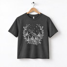 A black t-shirt with a white graphic design depicting a mountain landscape with a wreath of flowers and stars above it.