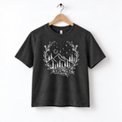 A black t-shirt with a white graphic design depicting a mountain landscape with trees, flowers, and stars.