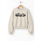 A beige sweatshirt with a graphic design of mountains and trees printed on it, hanging on a wooden hanger.