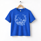 A blue t-shirt with a white graphic design depicting a mountain landscape with trees, flowers, and stars.