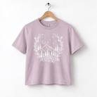 A light purple t-shirt with a white graphic design depicting a mountain landscape with trees, flowers, and stars.