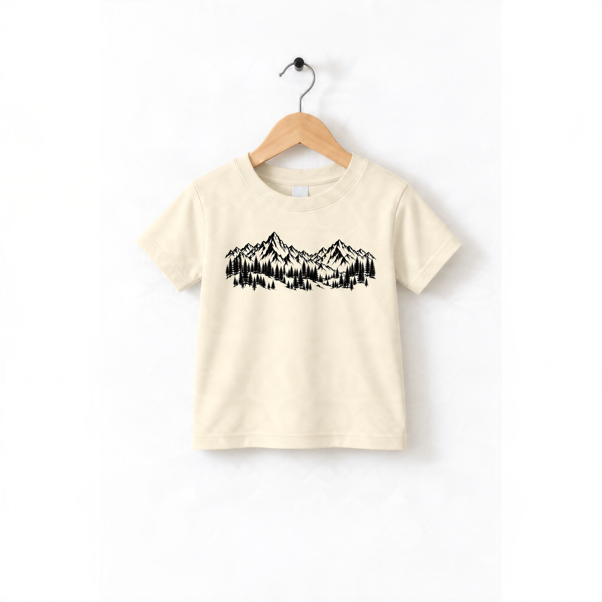 A beige t-shirt with a graphic design of mountains and trees printed on it, hanging on a wooden hanger.