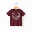 A burgundy-colored t-shirt with a white graphic design depicting a mountain landscape with trees, mountains, and stars.