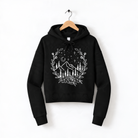 A black hoodie with a white graphic design depicting a mountain landscape with trees, flowers, and stars on a wooden hanger.