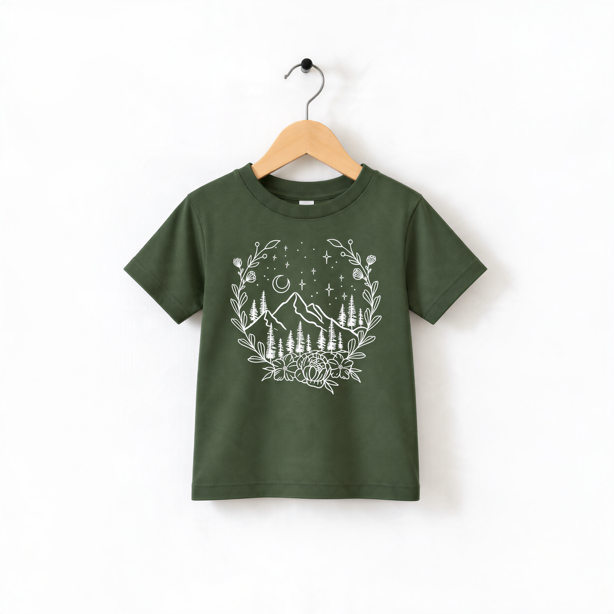 A green t-shirt with a white graphic design depicting a wreath of trees and mountains, along with stars and a crescent moon, hanging on a wooden hanger.