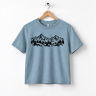 A light blue t-shirt with a graphic design of mountains and trees printed on it, hanging on a wooden hanger.