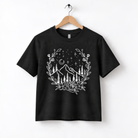 A black t-shirt with a white graphic design depicting a mountain landscape with trees, flowers, and stars in the night sky.