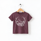 A burgundy-colored t-shirt with a white graphic design depicting a mountain landscape with trees, flowers, and stars.