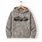 A camouflage-patterned hoodie with a graphic of mountains and trees printed on it, hanging on a wooden hanger.