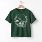 A green t-shirt with a white graphic design depicting a mountain landscape with trees, flowers, and a crescent moon.