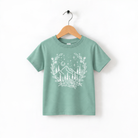 A green t-shirt with a white graphic design depicting a wreath of trees and mountains, along with stars and a crescent moon, hanging on a wooden hanger.