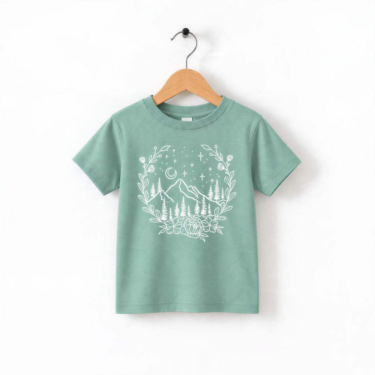 A green t-shirt with a white graphic design depicting a wreath of trees and mountains, along with stars and a crescent moon, hanging on a wooden hanger.