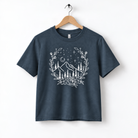 A navy blue t-shirt with a white graphic design depicting a mountain landscape with trees, flowers, and stars.
