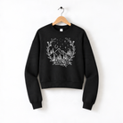 A black sweatshirt with a white graphic design depicting a wreath of flowers and trees, along with stars and mountains, hanging on a wooden hanger.