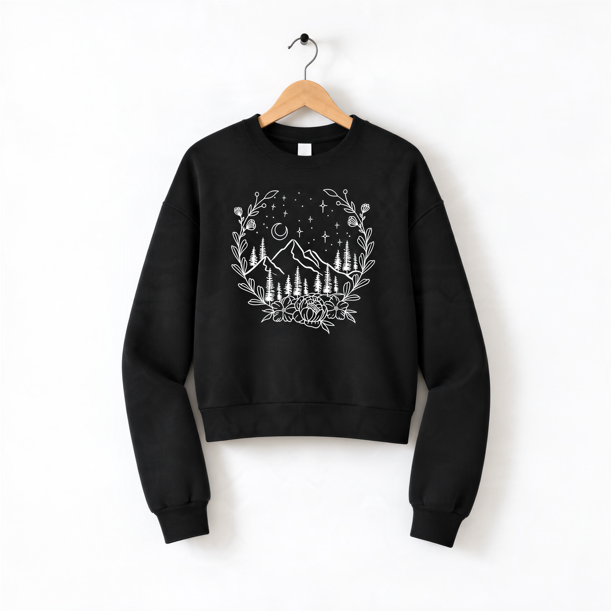 A black sweatshirt with a white graphic design depicting a wreath of flowers and trees, along with stars and mountains, hanging on a wooden hanger.