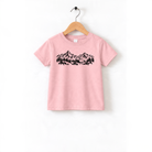 A pink t-shirt with a graphic design of mountains and trees printed on it, hanging on a wooden hanger.
