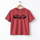 A red t-shirt with a graphic design of mountains and trees printed on it, hanging on a wooden hanger.