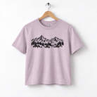 A light pink t-shirt with a graphic design of mountains and trees printed on it, hanging on a wooden hanger.