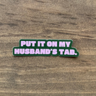 Sticker with text 'Put it on my husband's tab.' on a wooden surface