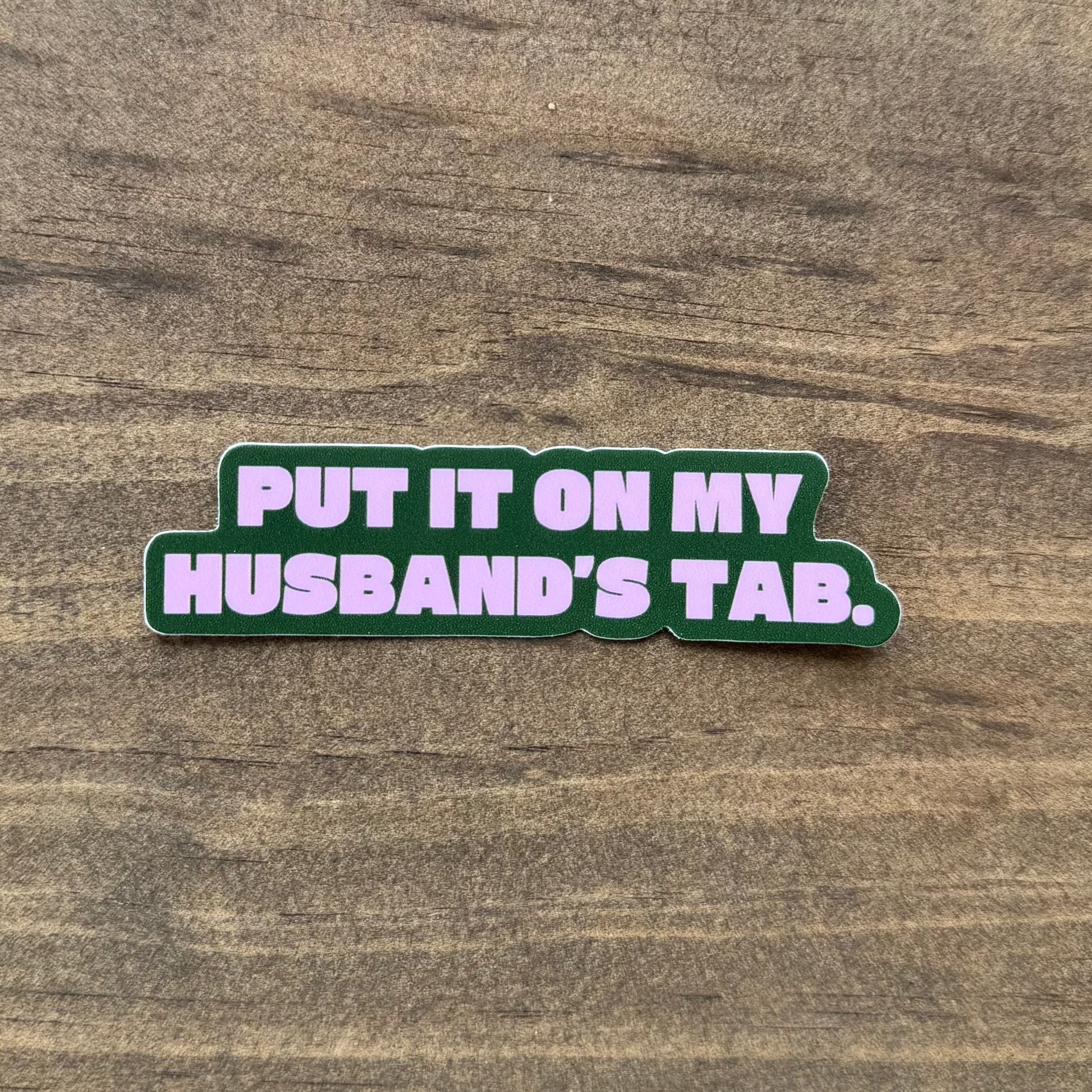 Sticker with text 'Put it on my husband's tab.' on a wooden surface