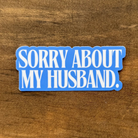 Blue sticker with text 'SORRY ABOUT MY HUSBAND.' on a wooden surface