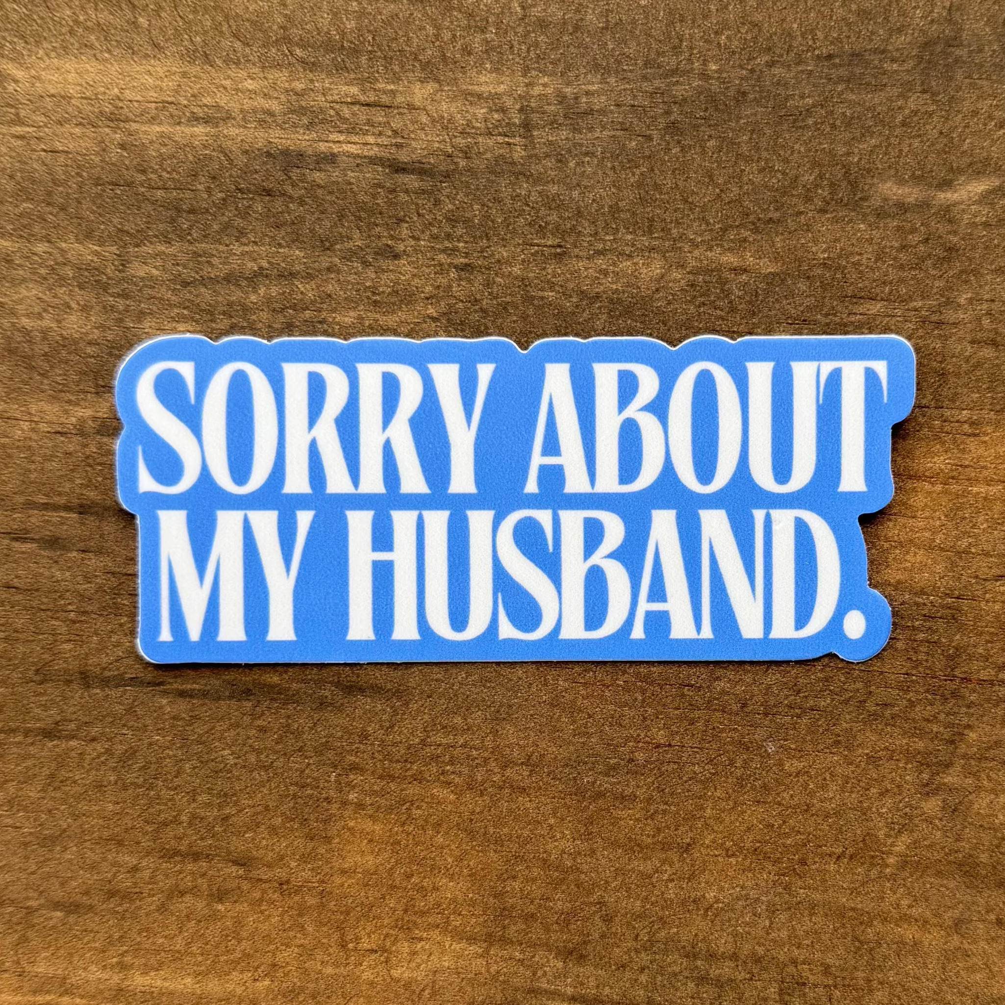 Blue sticker with text 'SORRY ABOUT MY HUSBAND.' on a wooden surface
