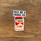 Sticker of a Coca-Cola can with text 'Pass Me a Fridge Cig' on a wooden surface