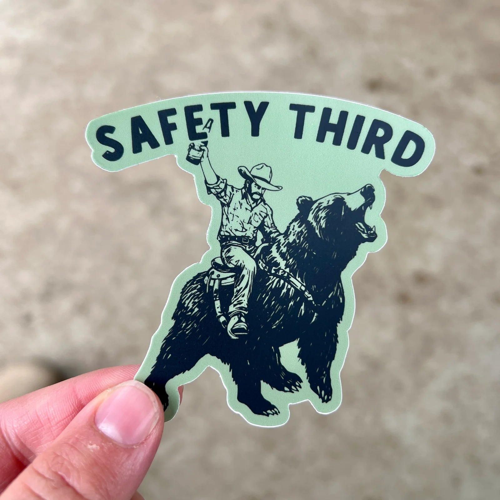Sticker with a cowboy riding a bear and 'Safety Third' text held by a hand against a neutral background