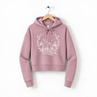 A pink hoodie with a white graphic design depicting a wreath of trees and mountains, hanging on a wooden hanger against a white background.