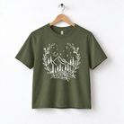 A green t-shirt with a white graphic design depicting a mountain landscape with trees, flowers, and stars in the night sky.