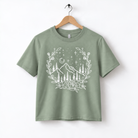 A green t-shirt with a white graphic design depicting a mountain landscape with trees, flowers, and stars.