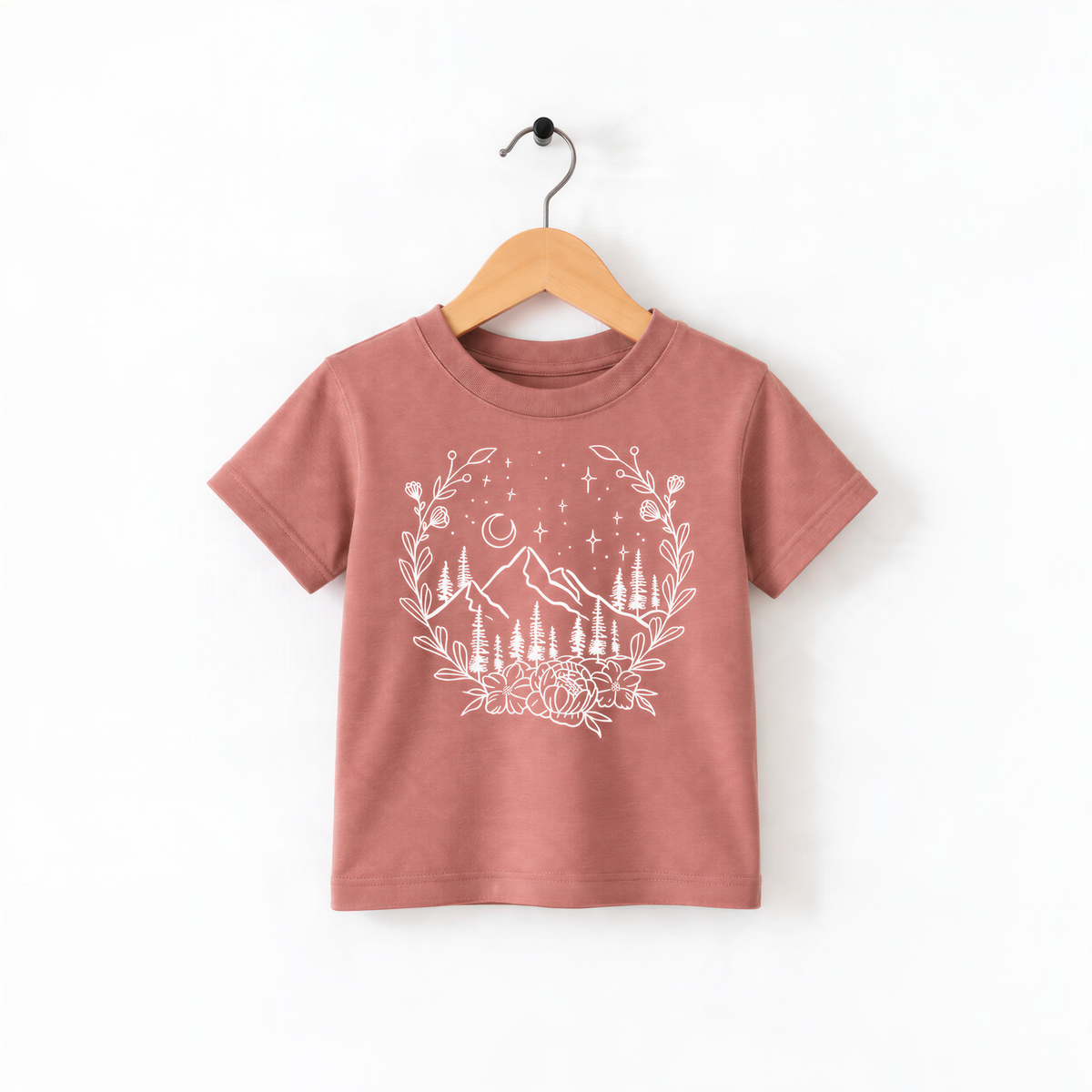 A pink t-shirt with a white graphic design depicting a mountain landscape with trees, flowers, and stars hanging from a wooden hanger.