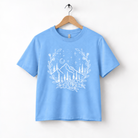 A light blue t-shirt with a white graphic design depicting a mountain landscape with trees, flowers, and stars.