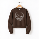 A brown sweatshirt with a white floral design on the front, hanging on a wooden hanger against a white background.