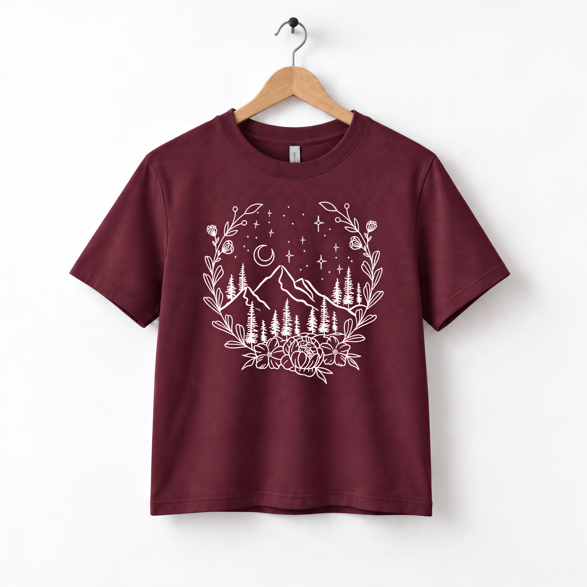 A burgundy-colored t-shirt with a white graphic design depicting a mountain landscape with trees, flowers, and stars.
