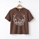 A brown t-shirt with a white graphic design depicting a mountain landscape with trees, flowers, and stars in the night sky.