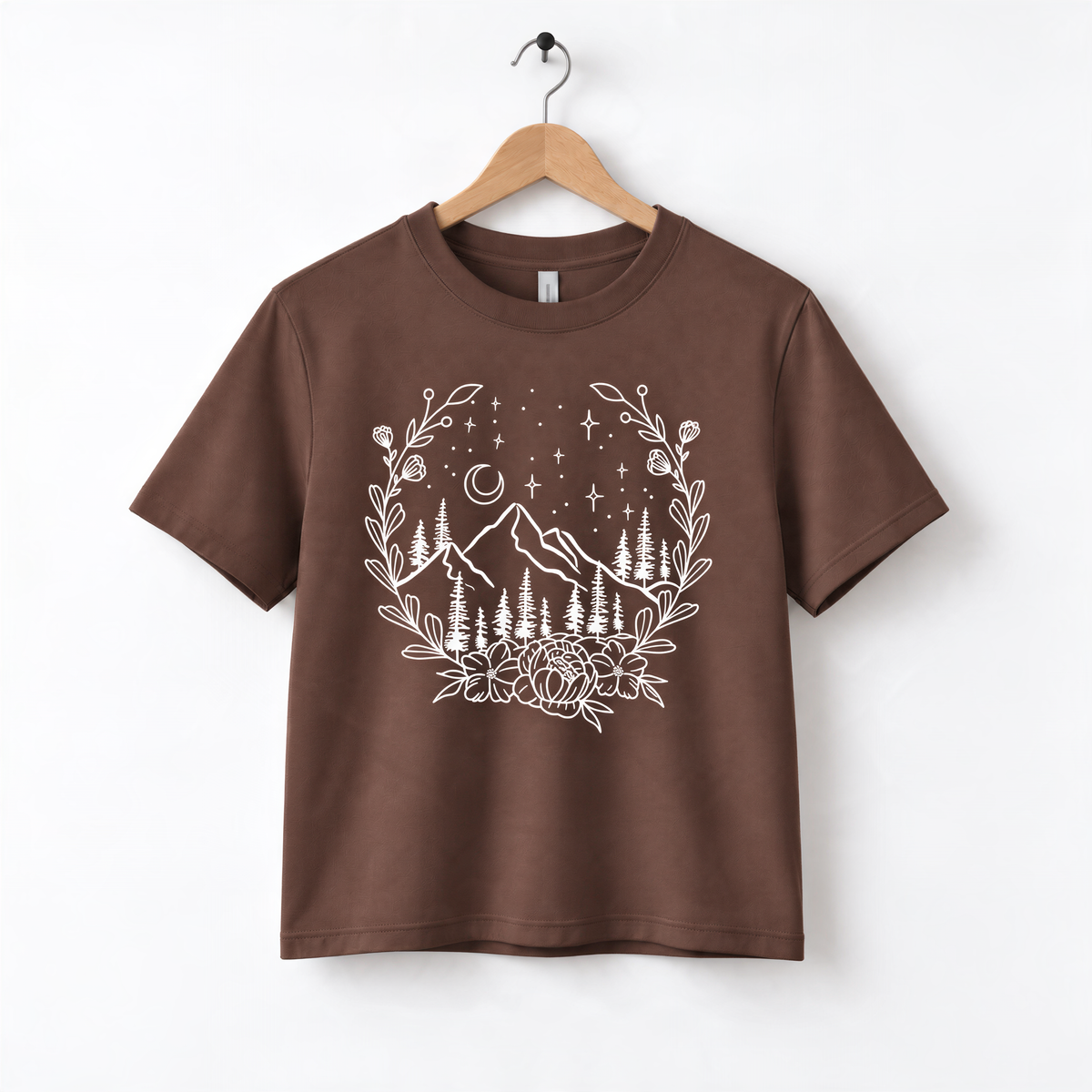 A brown t-shirt with a white graphic design depicting a mountain landscape with trees, flowers, and stars in the night sky.