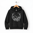 A black hoodie with a white graphic design depicting a wreath of trees and mountains, along with stars and a crescent moon, hanging on a wooden hanger.