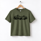 A green t-shirt with a graphic design of mountains and trees printed on it, hanging on a wooden hanger.