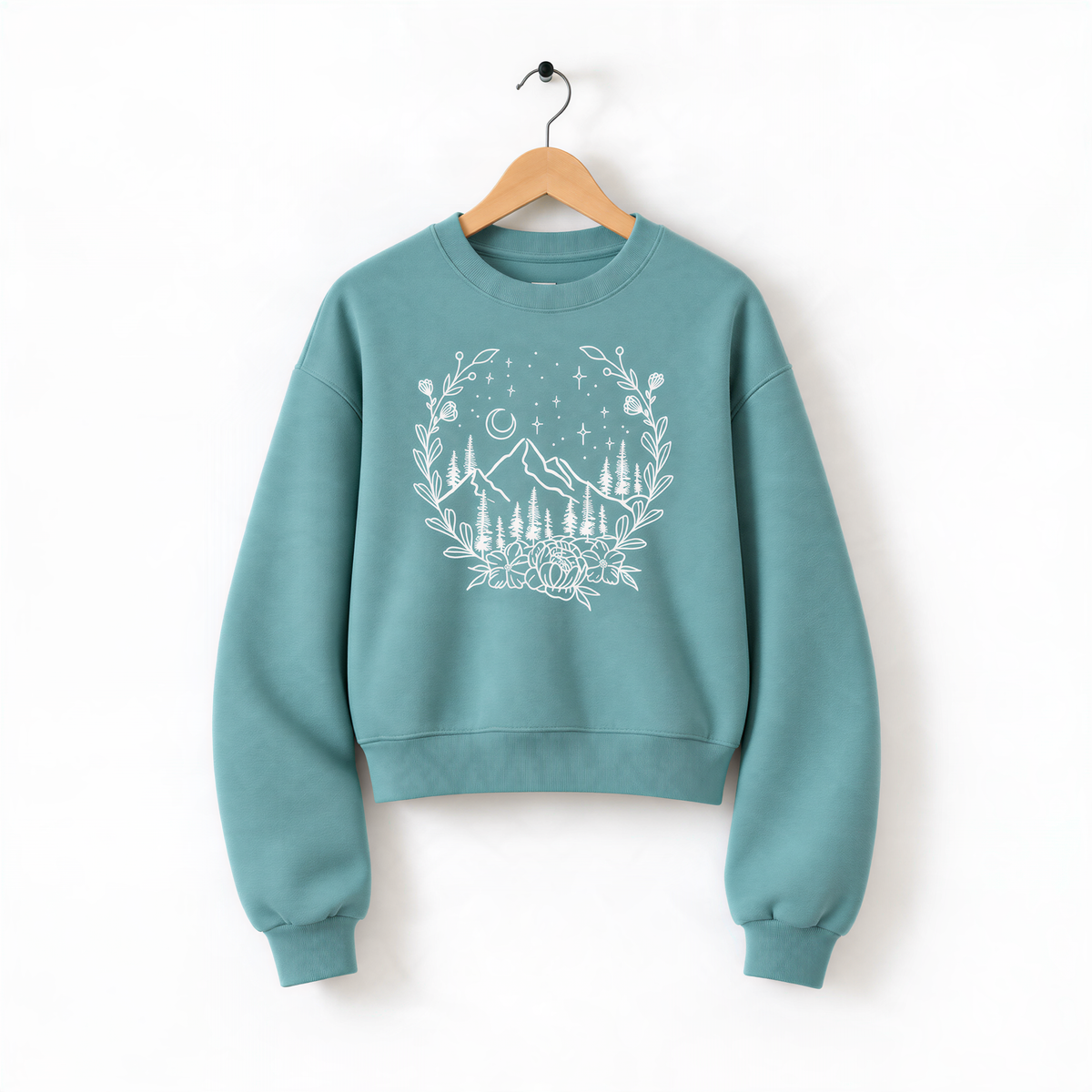 A teal-colored sweatshirt with a white graphic design depicting a wreath of trees and mountains, along with stars and a crescent moon, hanging on a wooden hanger.