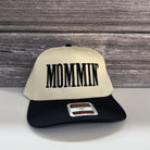 A white baseball cap with the word "MOMMIN" printed on it, placed against a wooden background.