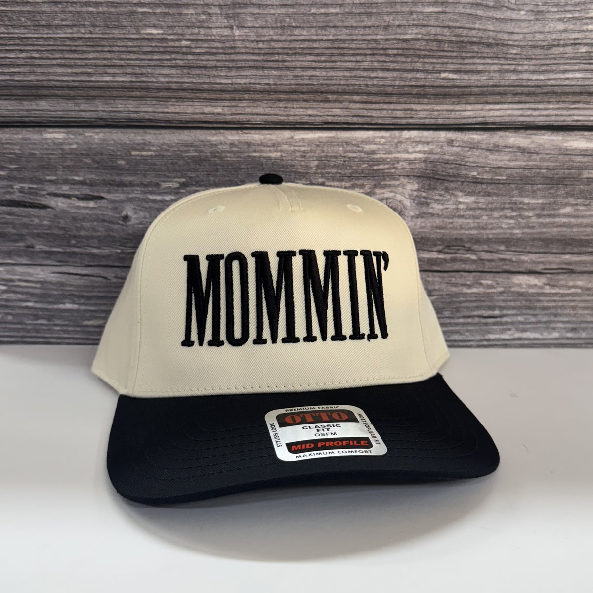 A white baseball cap with the word "MOMMIN" printed on it, placed against a wooden background.