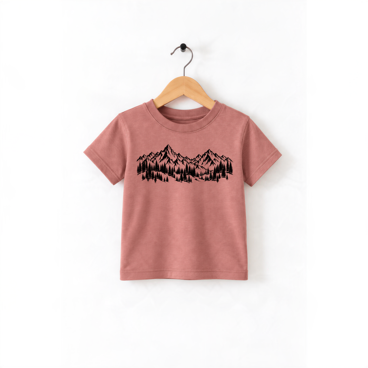 A pink t-shirt with a graphic design of mountains and trees printed on it, hanging on a wooden hanger.